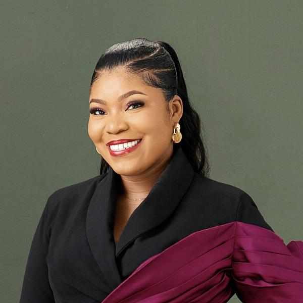 Headshot of Augustina Emele, Ashoka Africa staff. Person with tied back black hair about shoulder length and light brown skin smiling at the camera. Dressed in black business suit with a maroon sash. Background is green / grey