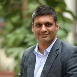 Photo of Yashveer Singh, Executive Director of Ashoka Young Changemakers. Person wiith short hair dressed in a suit smiling at the camera. Background of a garden plaza
