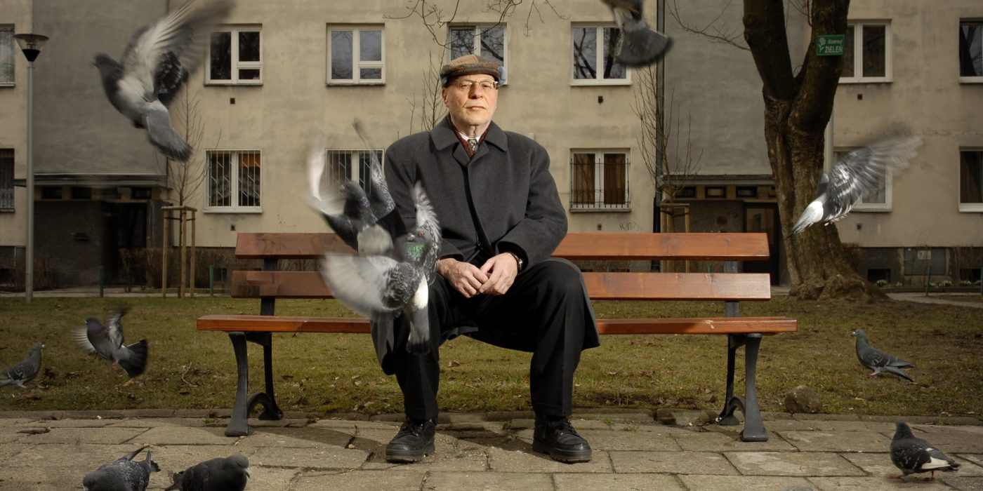 an older man in a cuff on a bench, pigeons flying nearby