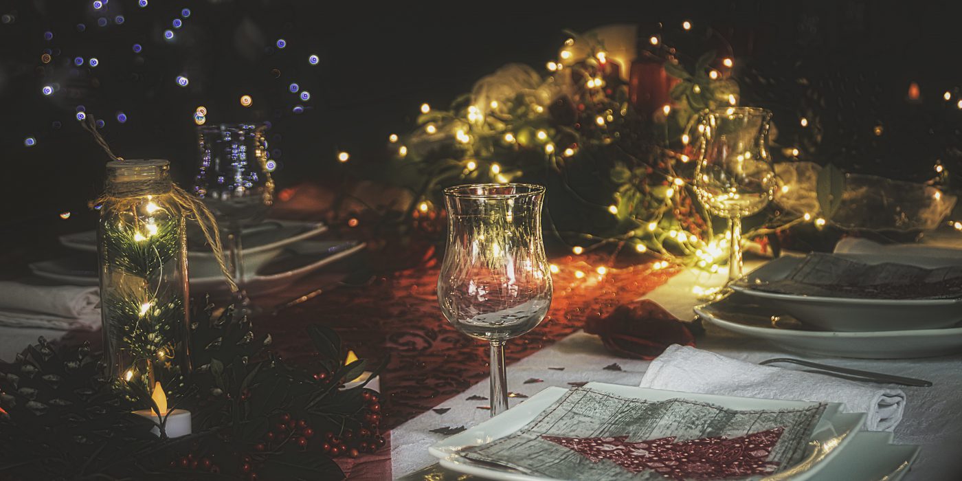 Zdjęcie pokazuje pięknie zastawiony świąteczny stół. Jest na nim dużo małych światełek, serwatki z choinkami i gałązki drzewa iglastego.