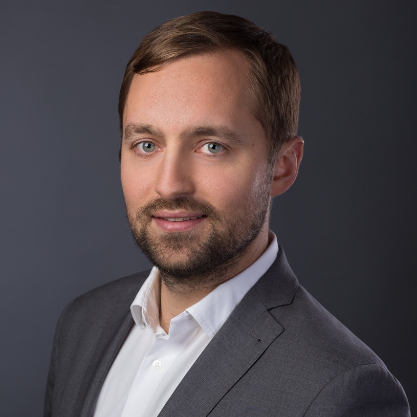 Portret Krzysztofa Arciszewskiego na szarym tle. Mężczyzna jest w garniturze, delikatnie się uśmiecha.
