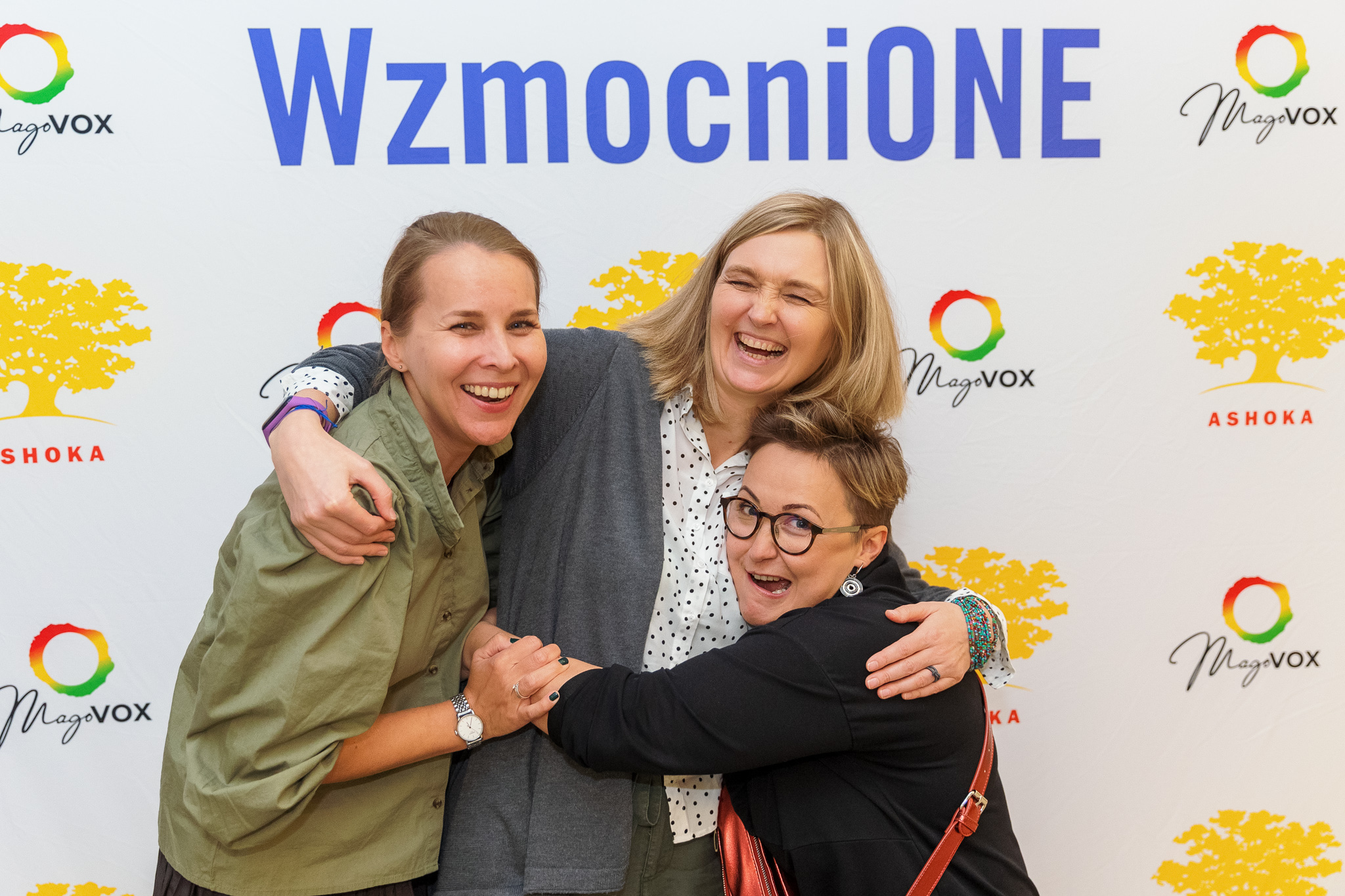 Zdjęcie przedstawia 3 członkinie Fundacji im. Julii Woykowskiej pozujące na białej ściance zdjęciowej z fioletowym napisem "WzmocniONE" na ich głowami oraz kolorowymi logami Ashoki i MagoVoxu. Kobiety przytulają się, śmieją się, jedna robi śmieszną wykrzywioną minę.