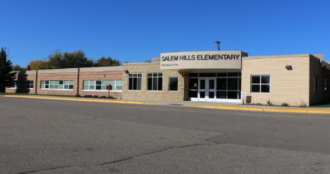 A long building with a tan half including a doorway and a sign above that reads "Salem Hills Elementary" with the left half made of brick and windows. The sky is blue and the asphalt in front of the building dark gray.