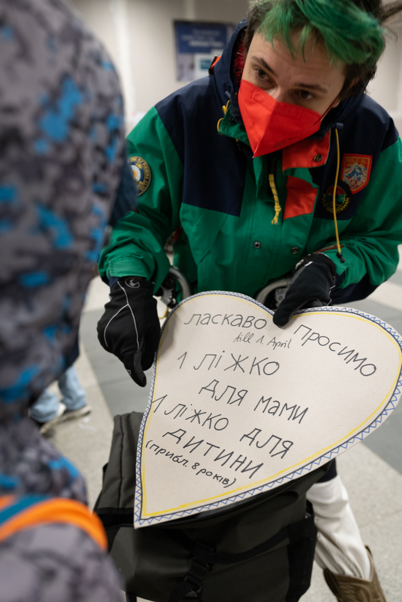 Person in a winter jacket with green hair with a red COVID mask on holding a poster up for another person to see. The poster is in the shape of a heart and has Ukrainian on it