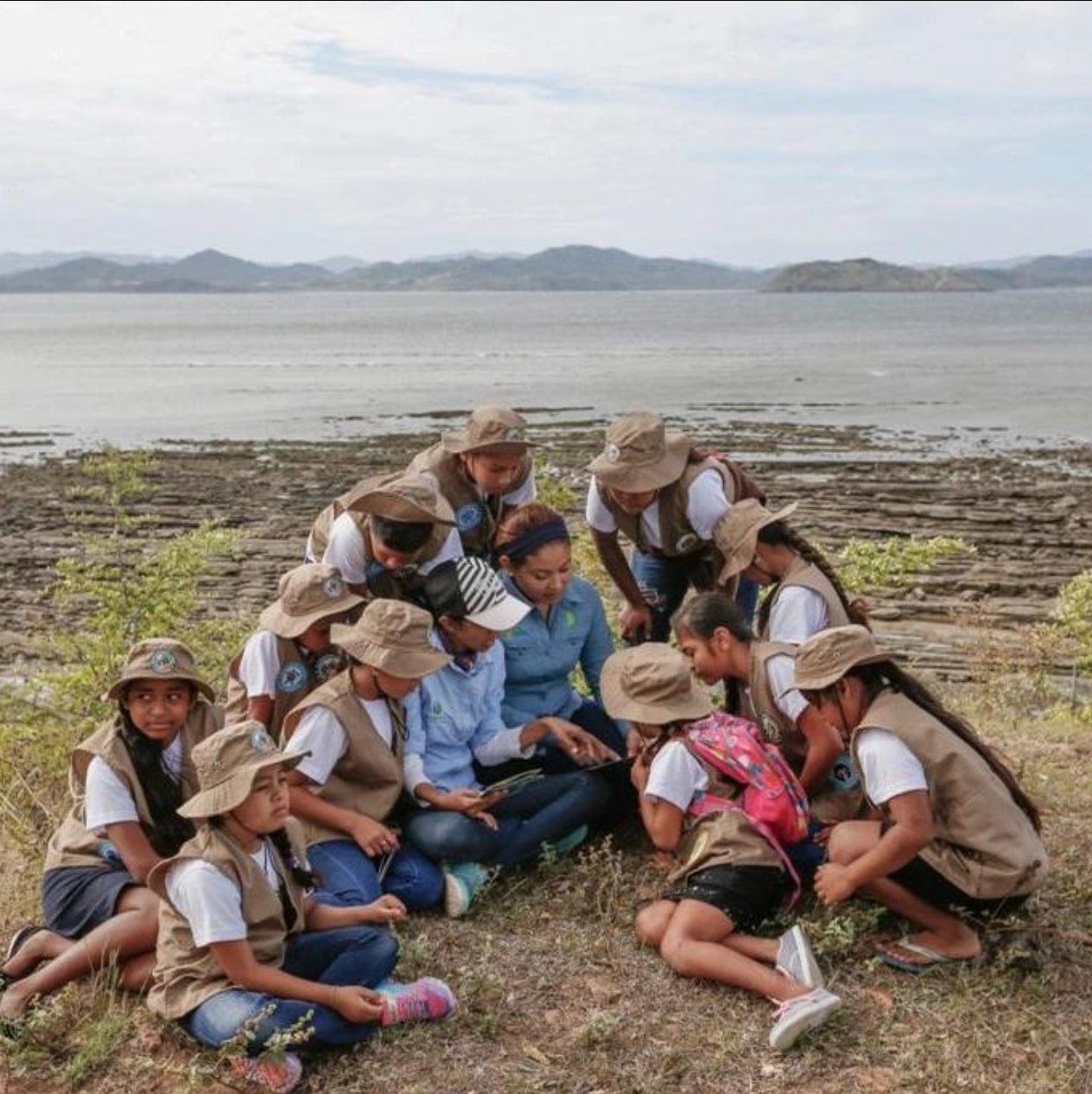 Una mujer que enseá a un grupo de niñas, niños y jóvenes. Se ve un espacio natural con lo que parece un lago y montañas de fondo. 