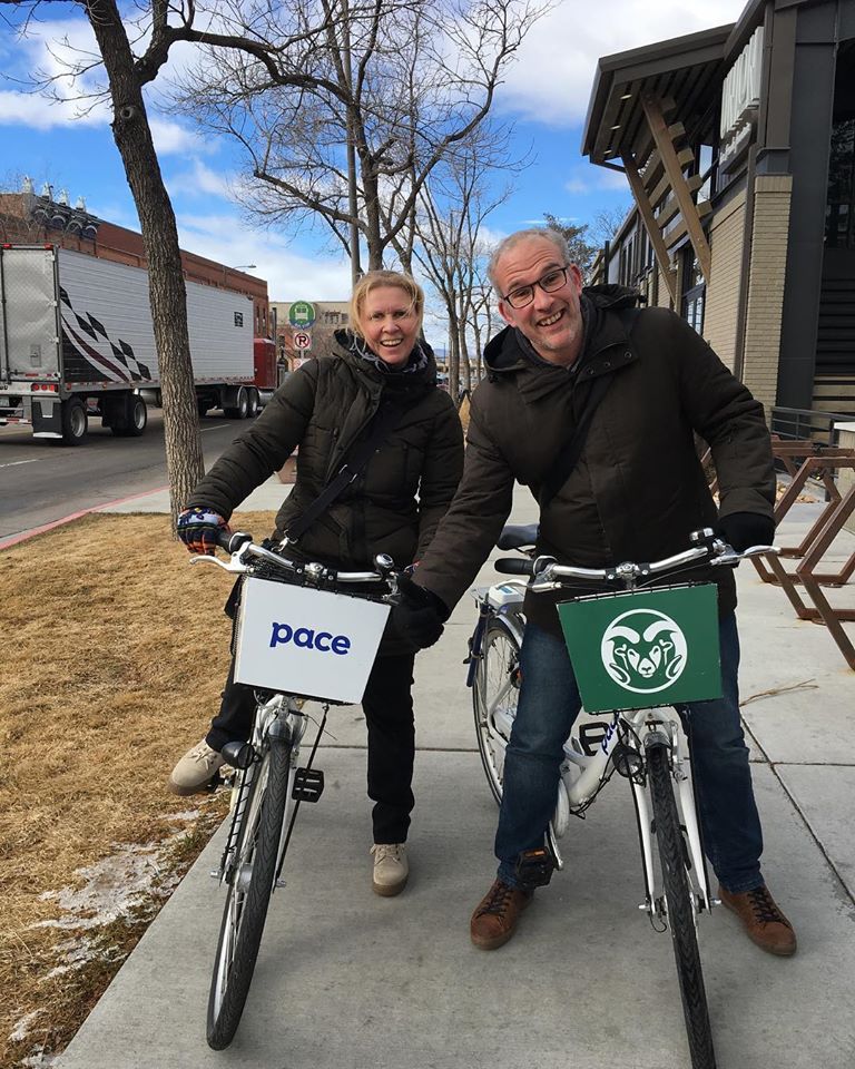 A man and a woman on their respective bikes, smiling. The man has grey hair and small eyes. The woman has light coloured hair and big eyes. They are both wearing dark coats.
