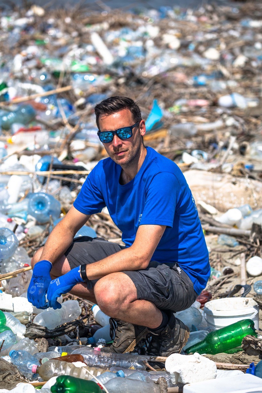 Man wearing glasses with short hair, wearing a blue t-shirt, blue gloves, and dark bermuda pants. He is surrounded by plastic.