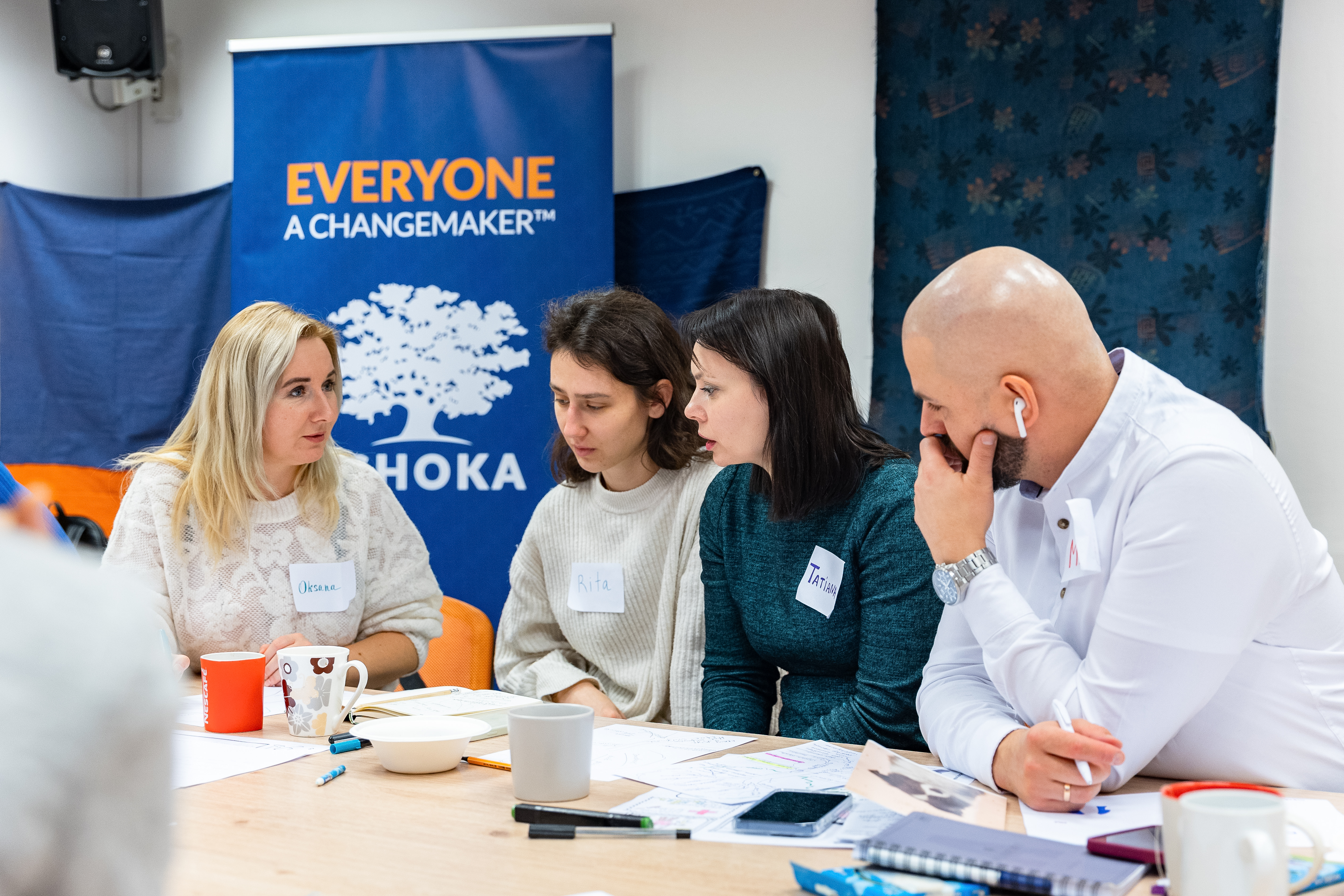 Zdjęcie przedstawia 3 kobiety i 1 mężczyznę rozmawiających przy stole. Za nimi widać rollup z napisem "Everyone a changemaker" i logiem Ashoki.