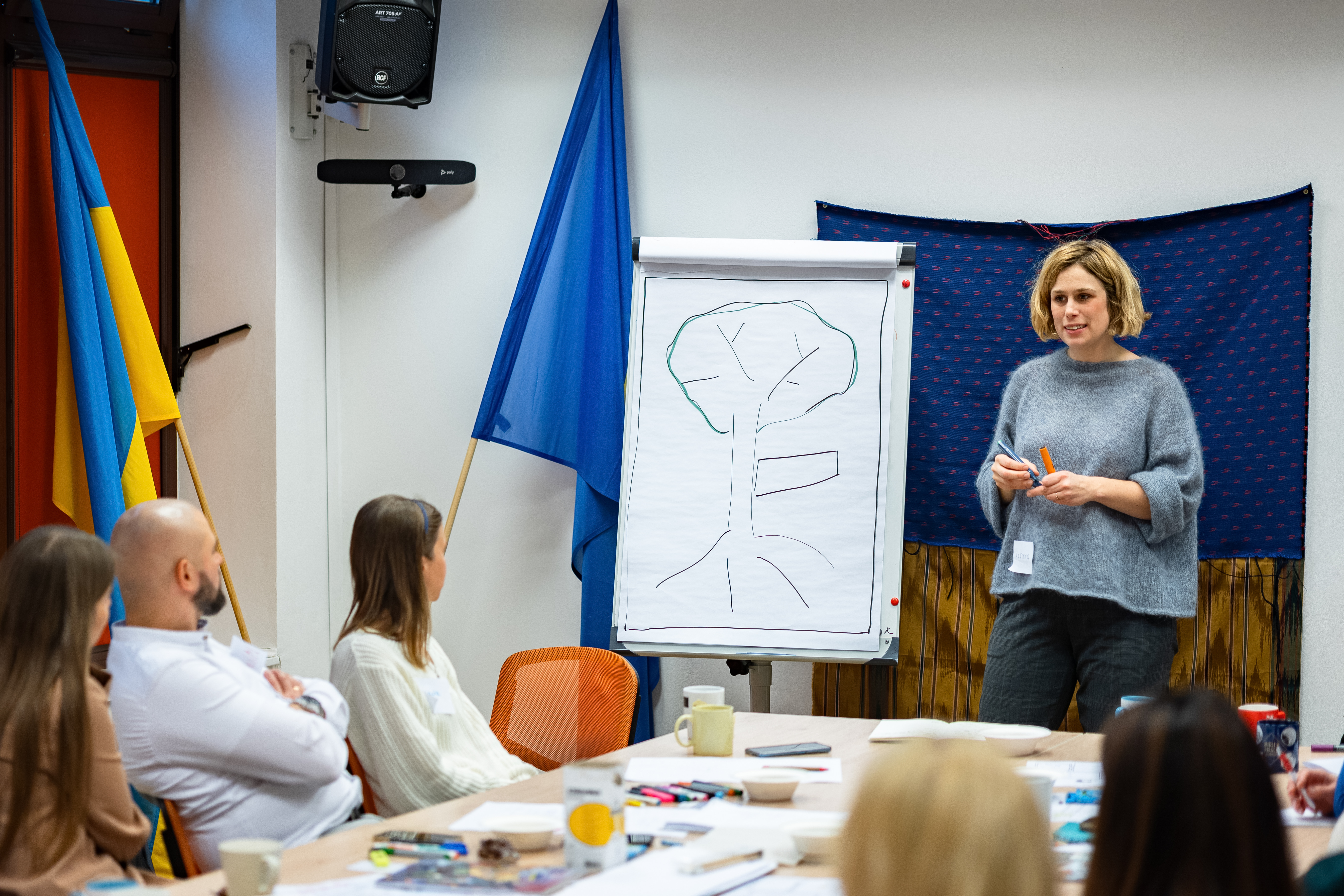 Zdjęcie przedstawia kobietę stojącą przy szczycie stołu, obok niej jest tablica z narysowanym drzewem i podpisem "Drzewo problemu". Za nią flaga Ukrainy i Unii Europejskiej. Przed nią przy stole siedzą i słuchają różne osoby.