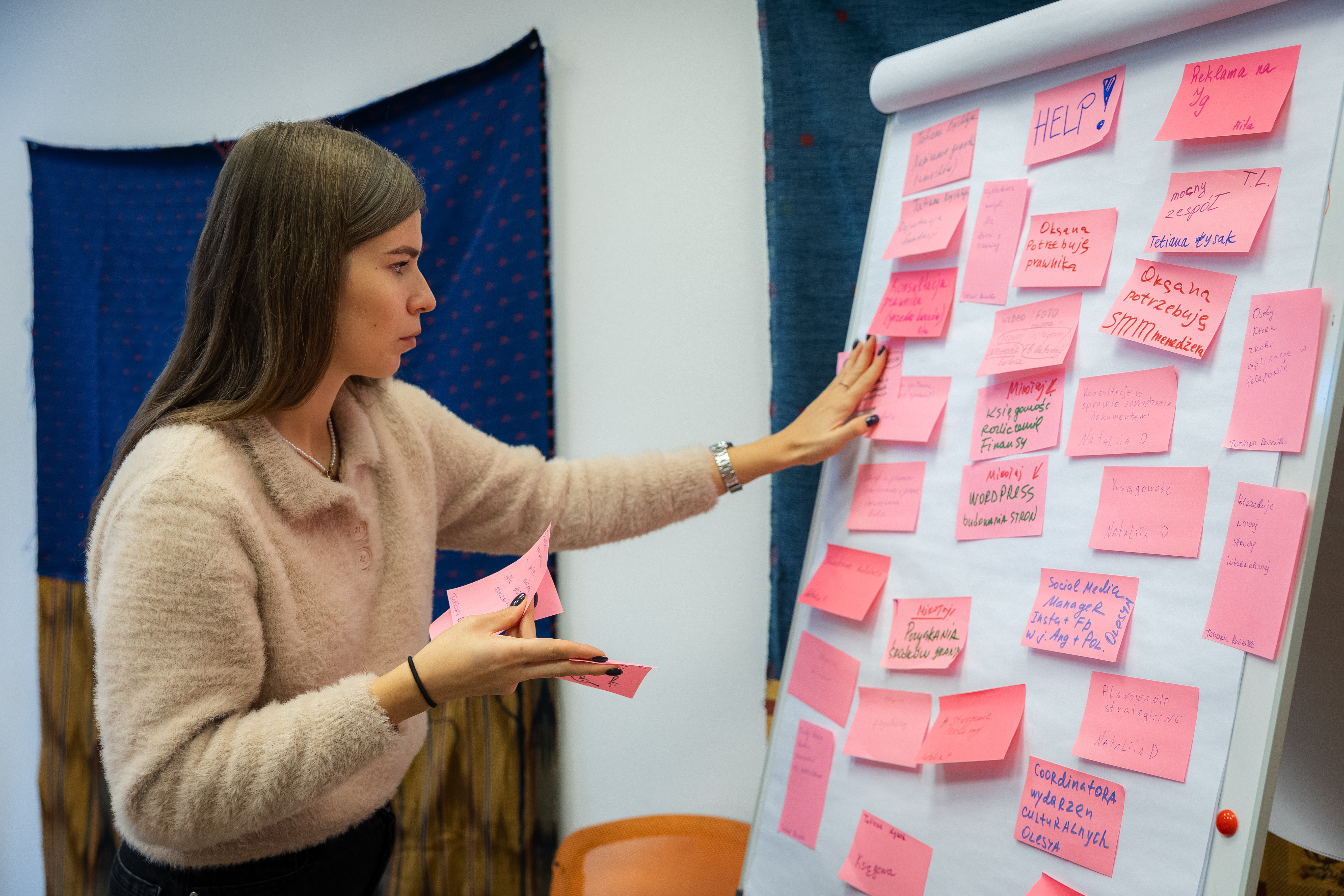 Zdjęcie przedstawia kobietę przyklejającą różową karteczkę post-it do białej tablicy, na której już wisi dużo takich samych karteczek z różnymi napisami.