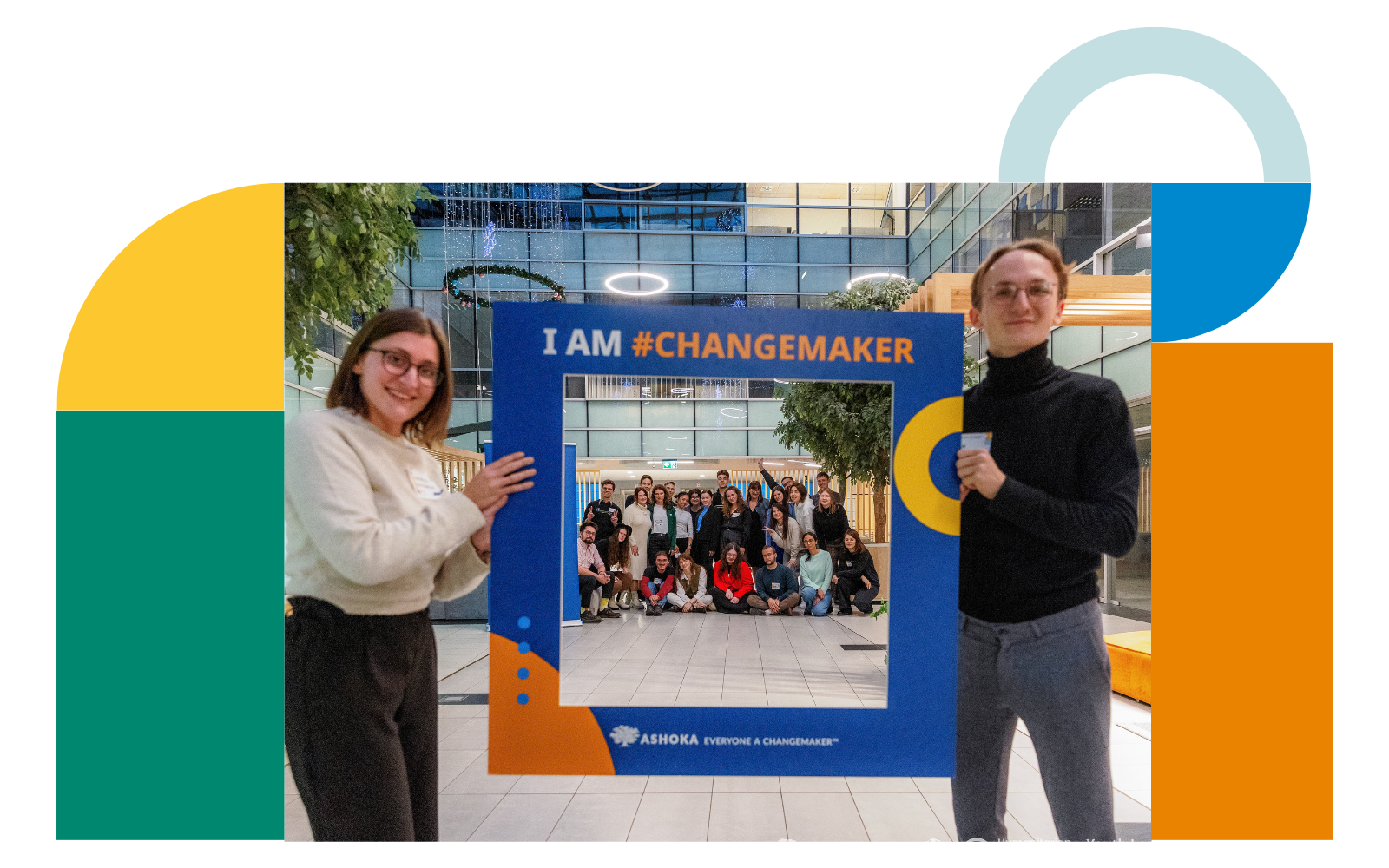 Two young people hold a cardboard frame reading 'I am #changemaker,' perfectly framing a group of smiling youths posing behind them.
