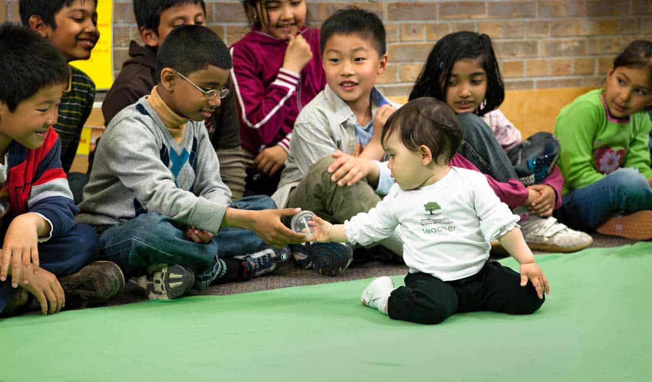 Baby handing an elementary school aged child an object while other students look on; located in a classroom