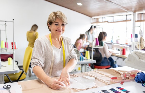 Person with shorter light hair and measuring tape across their shoulder measuring out fabric and smiling at the camera. Background of a fashion studio