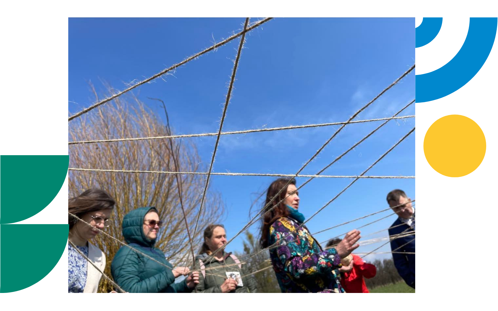 A group of people stands outdoors beneath a web of intersecting strings stretched between them. The activity takes place in an open, grassy area under a clear blue sky. The photo is framed by green, yellow, and blue geometric shape