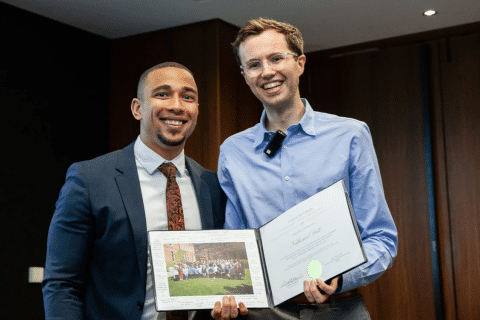 Ashoka Fellow Scott Stirrett (right, lighter skin glasses and shorter brown hair) with Nathaniel Scott (left, brown skin, dark short hair in a fade, goatee). Both people dressed in formal wear smiling at the camera. Background 