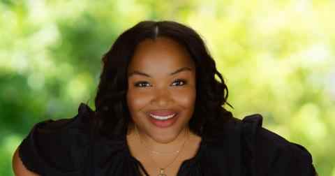 Headshot of Camesha Cox Canada Fellow 2023. Person with brown skin, long dark hair and lipstick smiling at the camera. Dressed in a black blouse; background of trees