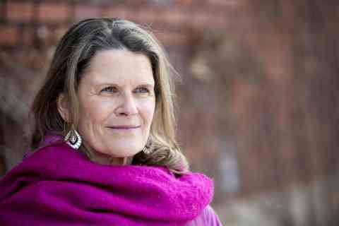 Photo of Canada Fellow Rachel Kiddell-Monroe. Photo of a person with longer brown hair and lighter skin smiling to the right of the camera. Dressed in a pink scarf; background of a brick wall.