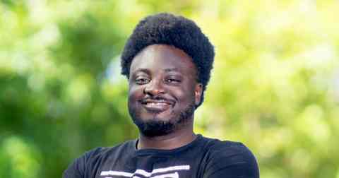 Headshot of Ernest Jr. Edmond. Person with dark brown skin and dark flat top smiling at the camera. dressed in dark t-shirt. Background of trees