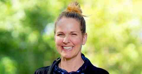 Headshot of Kat Kavanagh Canada Fellow 2023. Person with lighter skin, brown hair with blonde highlights in a bun smiling at the camera. Dressed in a dark jacket. Background of trees