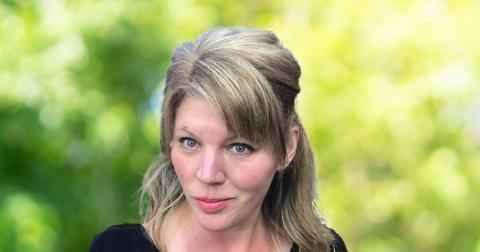 Headshot of Melanie Redman. Person with lighter skin and dark blonde hair smiling at the camera. Dressed in a dark black blouse. Background of trees