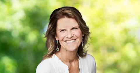 Headshot of Rachel Kiddell-Monroe Canada Fellow 2023. Person with lighter skin and shoulder-length dark brown hair with light brown highlights smiling at the camera. Dressed in a white blouse. Background of trees