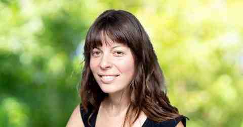 Headshot of Sara Dent Canda Fellow 2023. Person with lighter skin and shoulder-length dark brown hair with bangs smiling at the camera profile left. Dressed in a black blouse. Background of trees