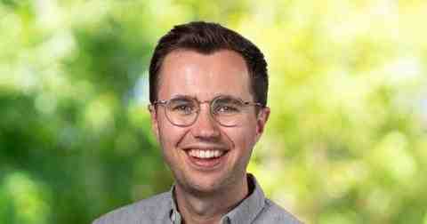 Headshot of Scott Stirrett Canada Fellow 2023. Person with lighter skin short dark brown hair and glasses smiling at the camera. Dressed in a light purple formal collard shirt. Background of trees