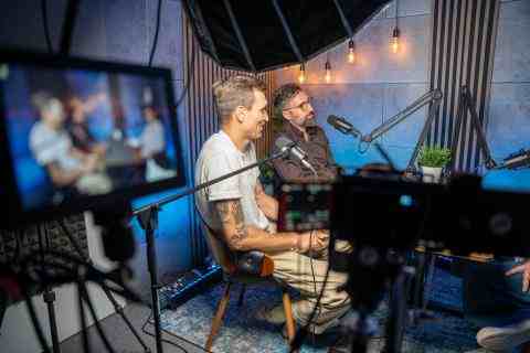 Two man sitting in podcast studio by the table talking to the microphone