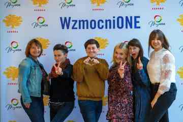 Na tle ścianki wzmocnionej z logo Wzmocnione, Ashoki oraz Magovox pozuje 6 osób w kolorowych ubraniach. Uśmiechają się, 3 pokazują znak peac/wolności palcami, jedna robi serduszko dłońmi.