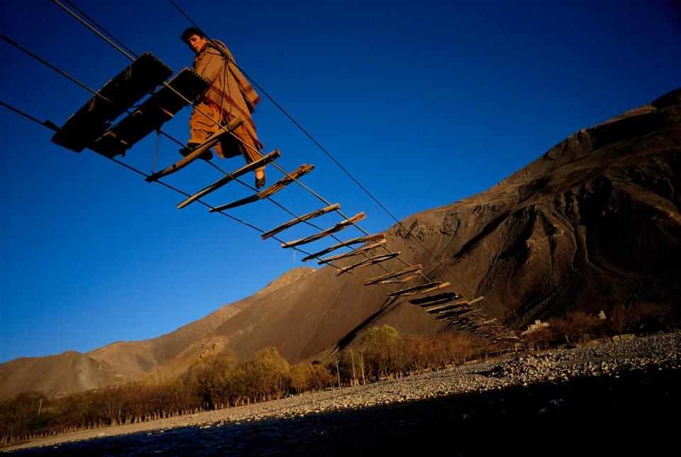 2020 Leading Social Entrepreneurs Front Cover. Depicting a man in the valleys of northeastern Afghanistan crossing a cable bridge with sparsely spaced wood planks above the Pandjchir River, connecting the village of Malespe to a refugee camp. Photographer and Ashoka Fellow, Reza Deghati, captured this shot in 2000.