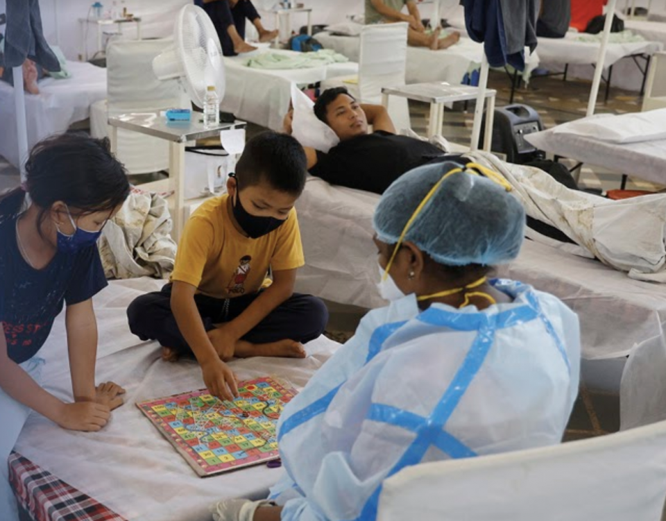 Picture of two young children playing a board game with a health care responder on a medical bed. Background of other people within a medical facility on or walking around medical beds.