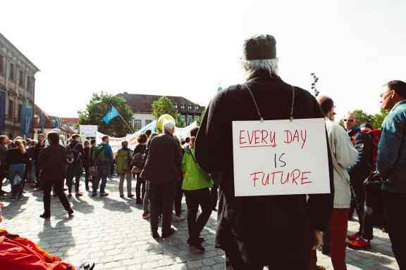 Mężczyzna w ciemnej kurtce ma na plecach zawieszony baner z napisem "every day is future". W tle widać innych protestujących,