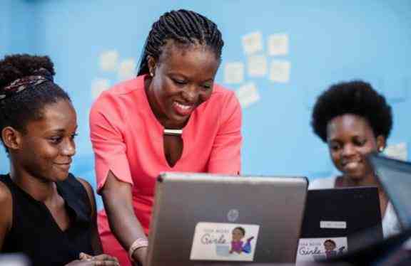 Na zdjęciu 3 kobiety wspólnie pracują przy laptopach. Uśmiechają się za nimi jasno-niebieska ściana.