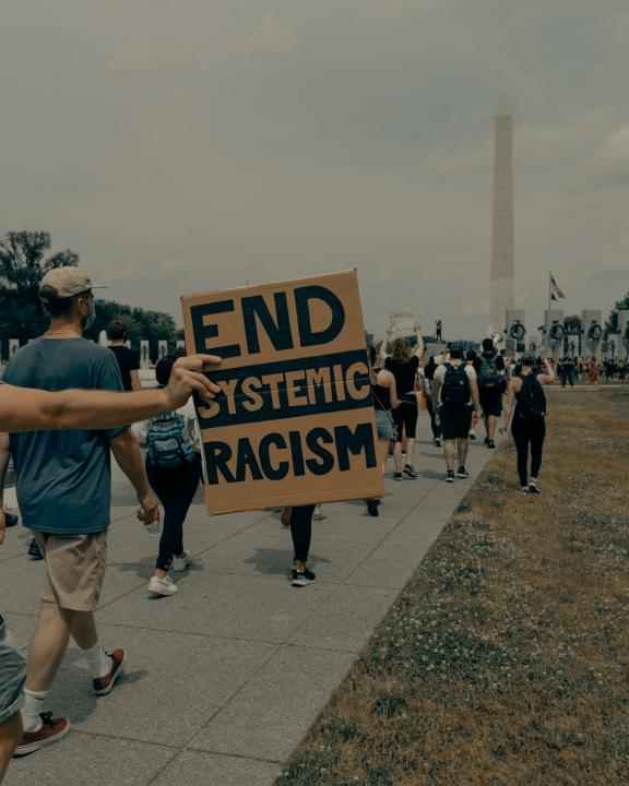 Karton z napisem "end systemic racism" (tłum. zakończmy systemowy rasizm) trzymana przez osobę protestującą. W tle inne osoby uczestniczące w proteście.