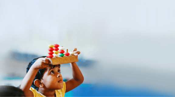 Criança vestindo uma camiseta amarela segura um ábaco sobre a cabeça e olha para o objeto, que possui cinco hastes verticais, que são preenchidas por bolinhas coloridas