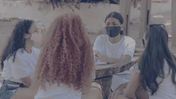 Grupo de 4 jovens mulheres conversando ao redor de uma mesa ao ar livre. Elas usam camiseta branca e máscara de proteção