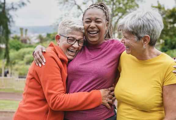 Três mulheres idosas riem e se abraçam em um parque
