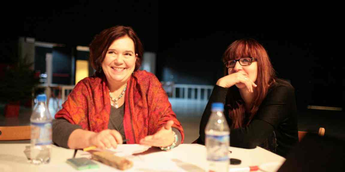 Zdjęcie przedstawia dwie uśmiechnięte kobiety siedzące przy stole. Po lewej Katarzyna Oleś w czerwonej chuście na ramionach i kolorowym naszyjniku.