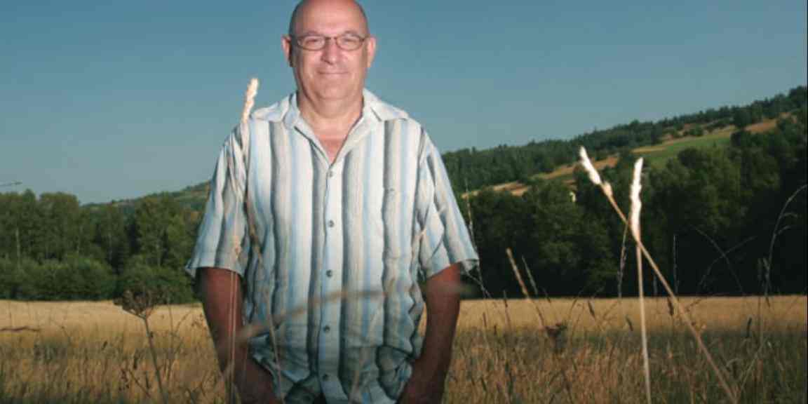 Zdjęcia Piotra Topińskiego na tle pola. Piotr ubrany jest w białą koszulę w pionowe pasy. Uśmiecha się.