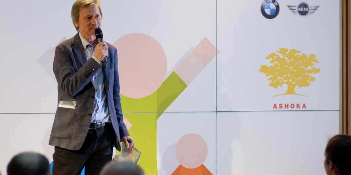 Zdjęcie Zdzisława Nitaka mówiącego do mikrofonu na scenie. W tle logo Ashoki.