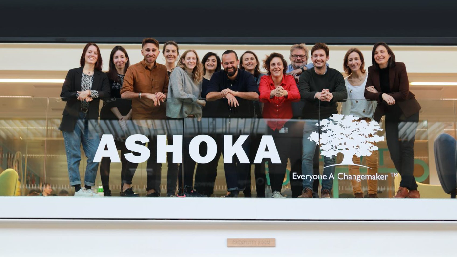 12 people smiling on a balcony with the word "Ashoka" below in white, and a white tree next to it