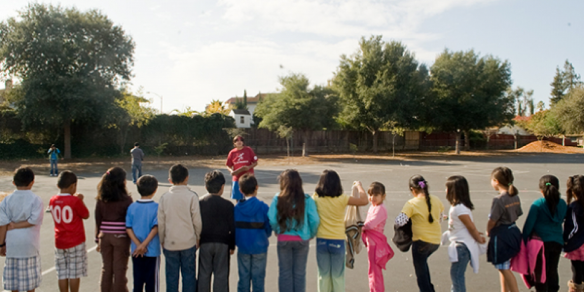 Zdjęcia przedstawia dzieci ustawione w rządku na szkolnym patio. Przed nimi stoi nauczyciel. Szykują się do zabawy.