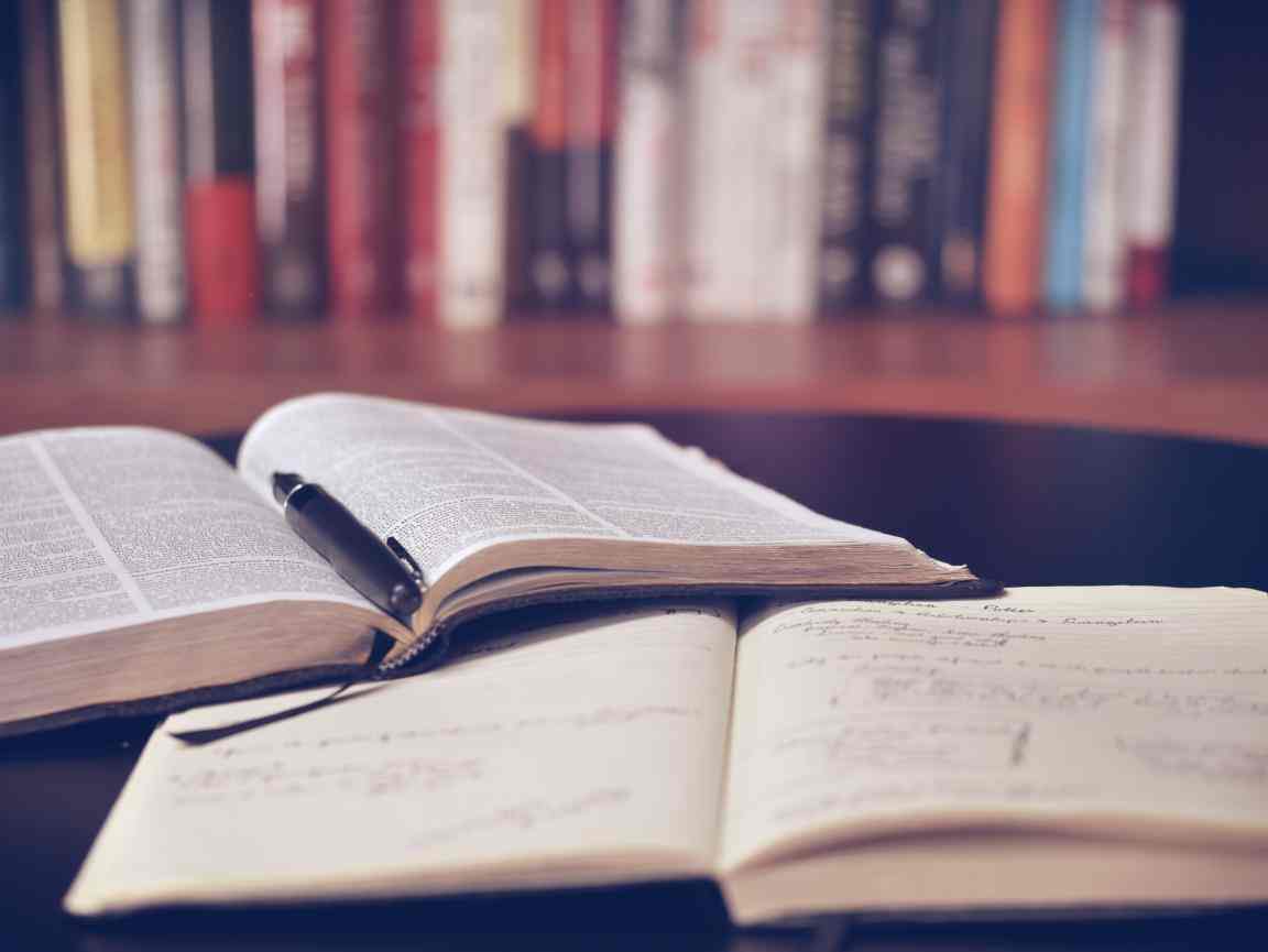 Na zdjęciu widać otwartą książkę i zeszyt oraz długopis leżące na stole. W tle półka z książkami.