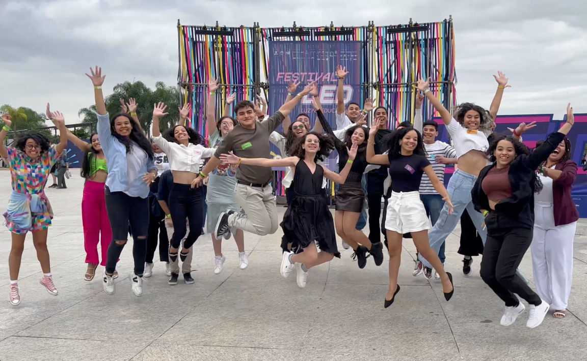 Grupo de jovens ao ar livre pulando. Ao fundo, há uma estrutura colorida com os dizeres "Festival LED"