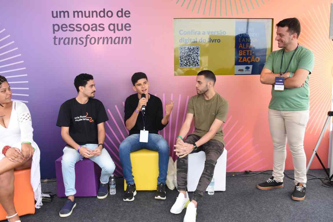 Quatro jovens sentam em pufes coloridos e um deles está em pé. Um deles fala ao microfone. O grupo está de frente a uma parede colorida, onde se lê "um mundo de pessoas que transformam"