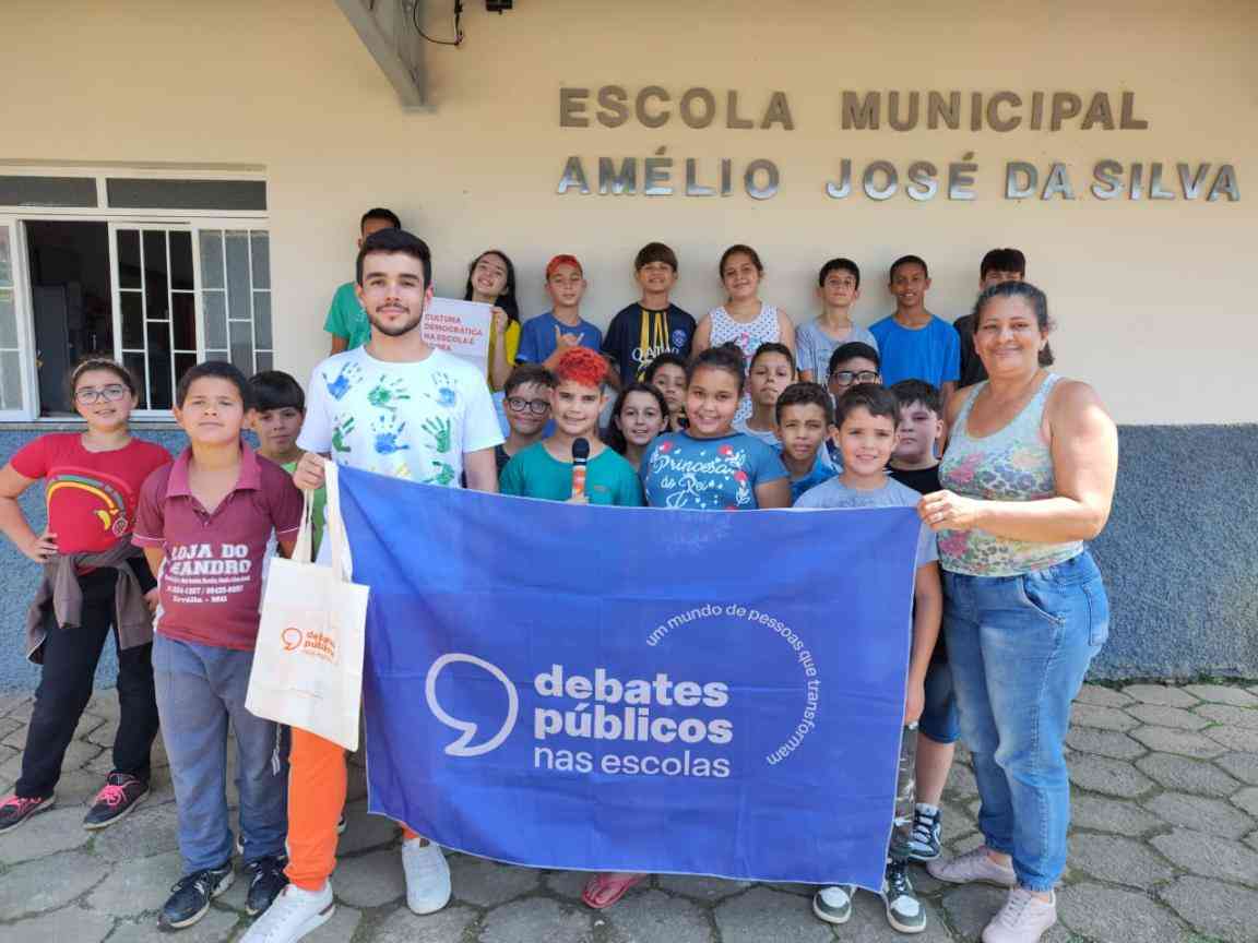 Grupo de crianças reunidas em frente à escola. Dois adultos estão junto com o grupo e seguram a bandeira dos Debates Públicos nas Escolas