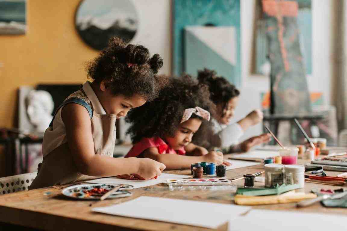 Duas crianças negras estão sentadas a uma mesa em que há tintas. Elas estão desenhando