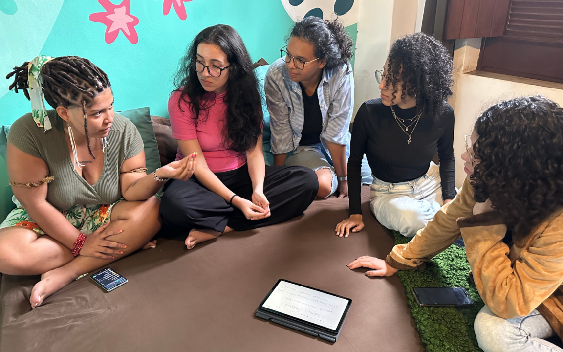 Vê-se 5 mulheres diversas sentadas em um semicírculo conversando