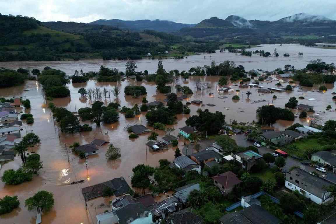 Imagem aérea mostra uma cidade atingida por enchentes, com a água em tom amarronzado chegando até o telhado das casas e copa das árvores. Ao fundo, vê-se montanhas