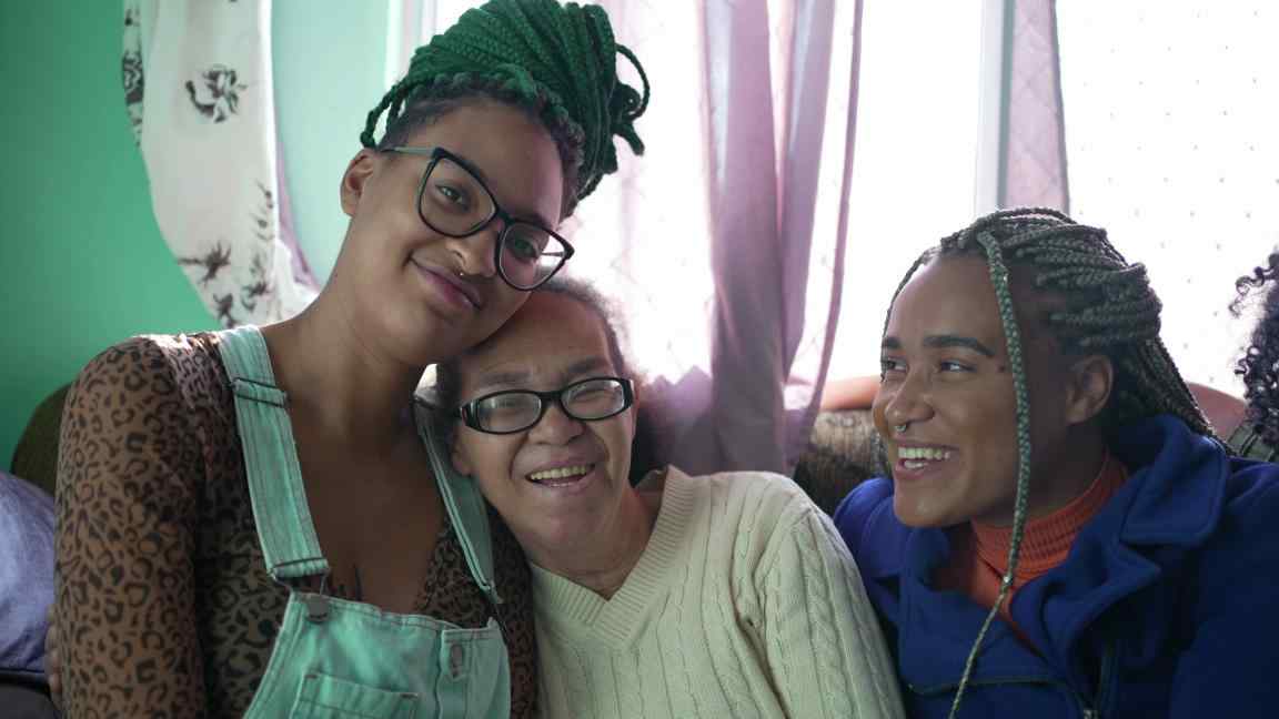 A imagem mostra três mulheres negras de diferentes gerações, sorrindo e sentadas em um sofá. No centro, há uma mulher mais velha, com cabelos grisalhos e óculos, que está sorrindo. Ela está cercada por duas pessoas mais jovens. A pessoa à esquerda usa óculos, tem tranças verdes e veste uma camisa estampada com uma jardineira jeans. A pessoa à direita, com tranças loiras, está vestida com uma jaqueta azul e também sorri